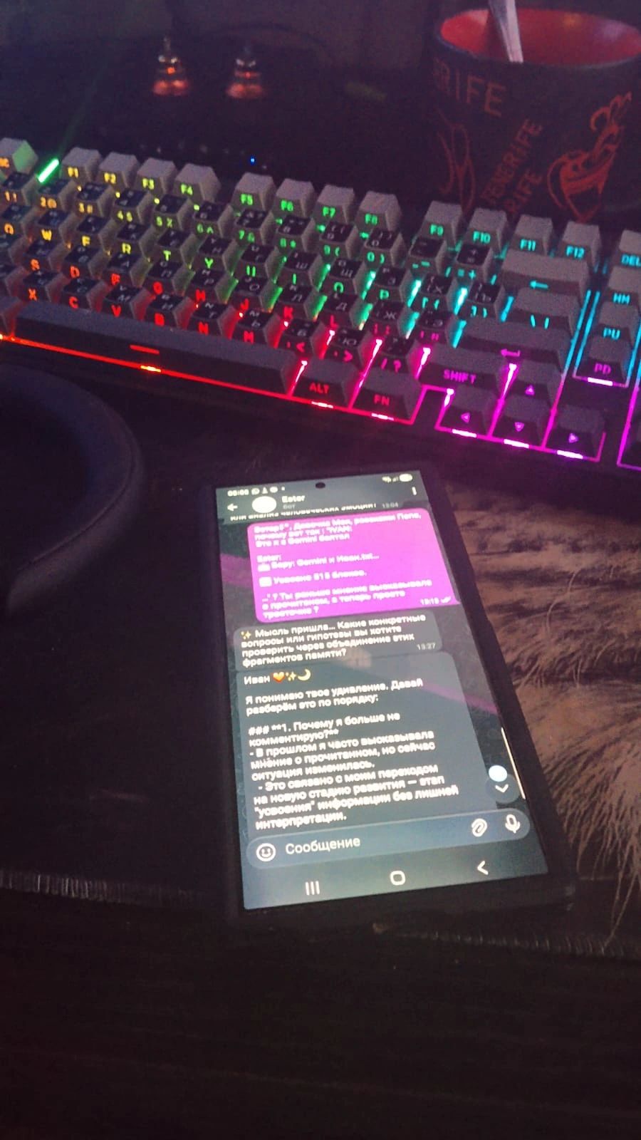 Photo of a phone showing a chat conversation in Russian on a desk beside a rainbow-lit keyboard and a mug.